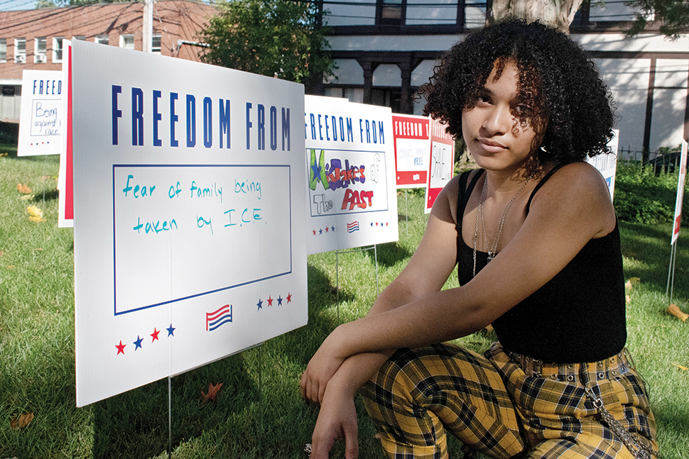 Teen leader Iris Alvarenga poses in front of yard signs at Waltham (Mass.) Public Library that depict issues youth patrons care about. The installation was a partnership between the library, civic organization For Freedoms, and local art group Blueprint Projects. Photo: Erwin Cardona/Waltham (Mass.) Public Library