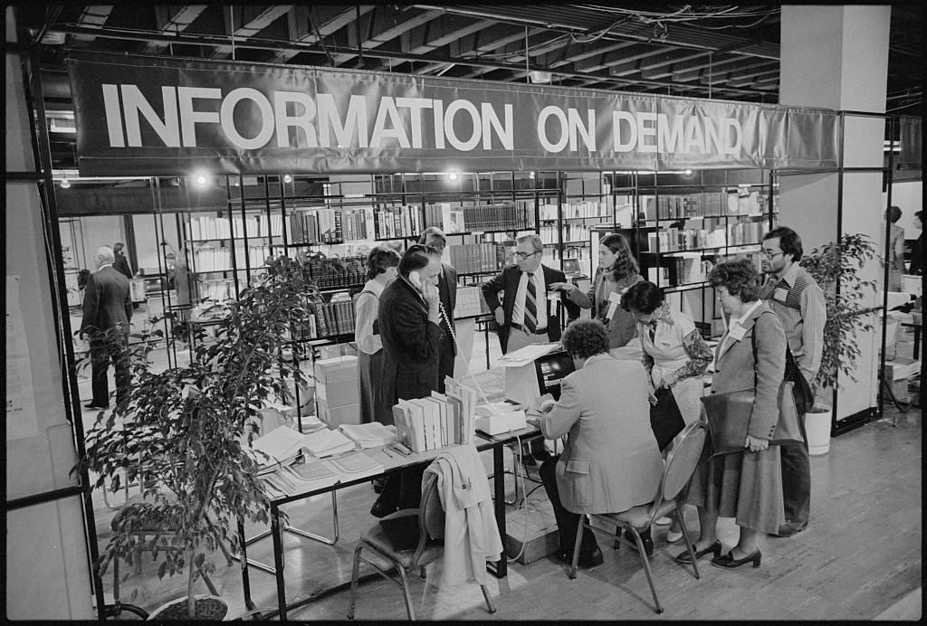 Black and white photograph of a man using computer as others watch at a exhibit with banner “Information on Demand”, at the White House Conference on Library and Information Services, Washington, D.C.