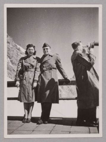 Man and Woman (Jimmy Ualade and Sylvia Mathon) standing together leaning against railing with mountain tops in the background. Additional man looking through the telescope next to them.