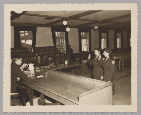 Empty courtroom except for two women, Sylvia and Sylvia, with their right hands raised to be sworn in to court. President of the court seated behind the desk. Four other men seated at a table next to the stands. 