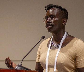 Bee Okelo, administrative and GIS analyst at San Francisco Public Library, is wearing a beige scoop neck t-shirt and glasses while speaking at a podium.