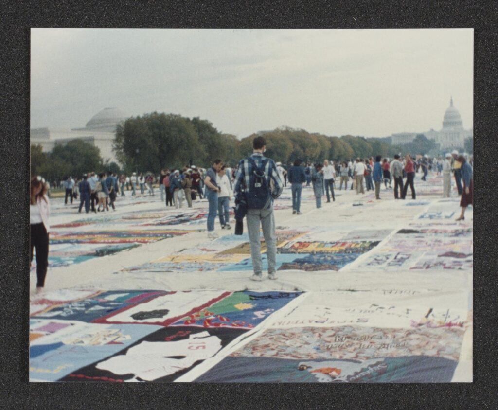 Color photograph of the AIDS quilt on the National Mall. The US capitol building is in the background. Foreground features visitors walking among the quilt.