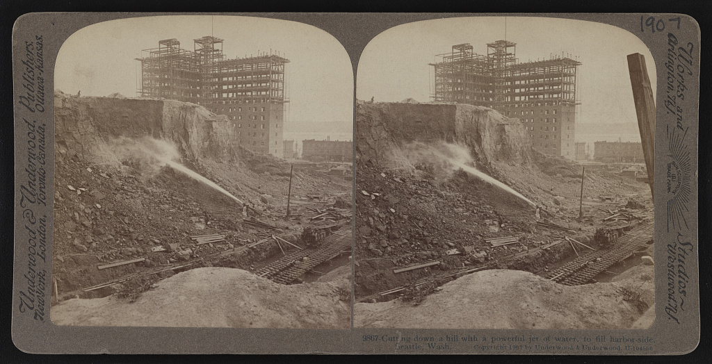 Stereograph showing water blasting into the side of a hill causing the dirt to crumble and slide downward; building under construction in background.