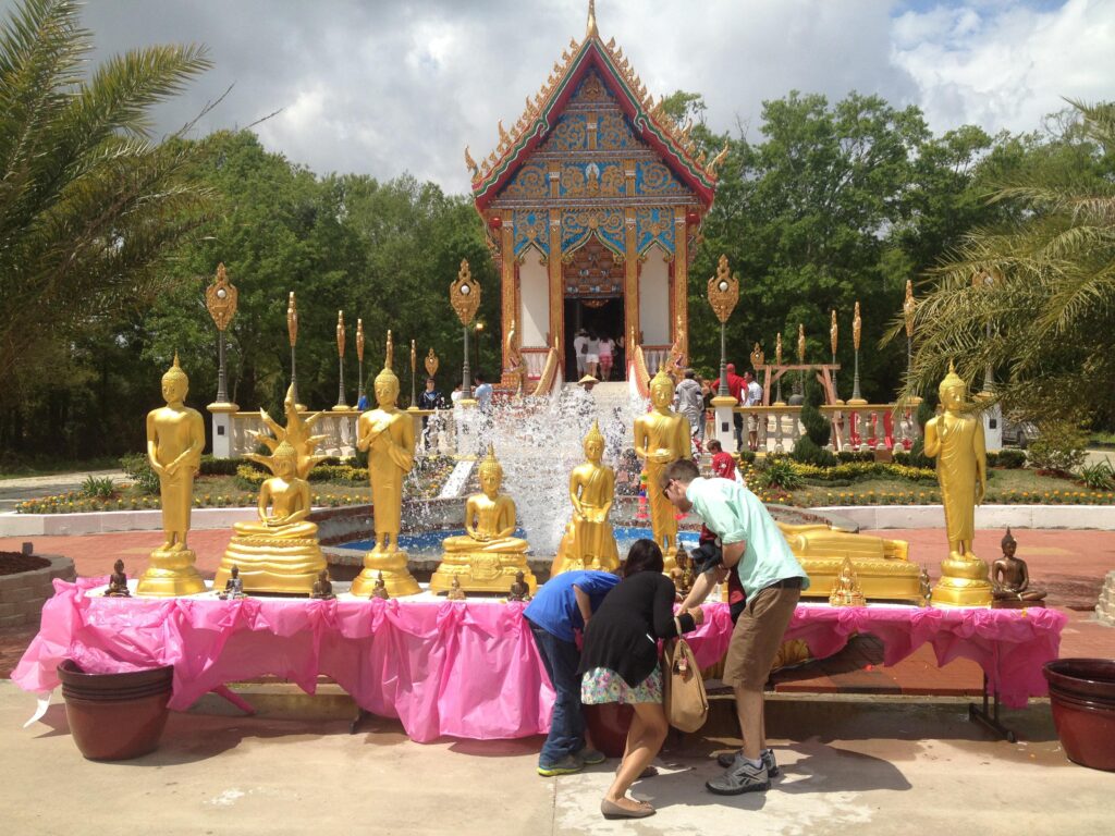 Three people crouching in front of an outdoor altar with a half dozen golden Buddhas with a large fountain behind them.